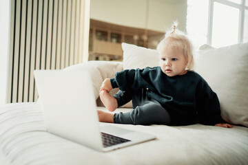 Cute little girl with blonde blue eyes. Children and the computer. watching an exciting children's program on a laptop on the Internet. Spends time at home on the couch. 