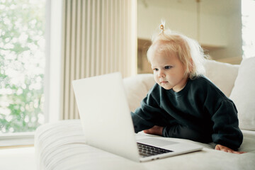 A child plays on a laptop computer. Portrait of a cute little girl with blonde blue eyes. A cheerful mood. Children and home. The family spends time at home on the couch.