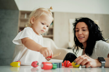 A babysitter and a child play with children's wooden toys in the apartment together. Portrait of a cute little blonde girl. A cheerful mood. Children and home. The family spends time at home.