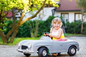 Little adorable toddler girl driving big vintage toy car and having fun with playing outdoors. Gorgeous happy healthy child enjoying warm summer day. Smiling stunning kid playing in domestic garden
