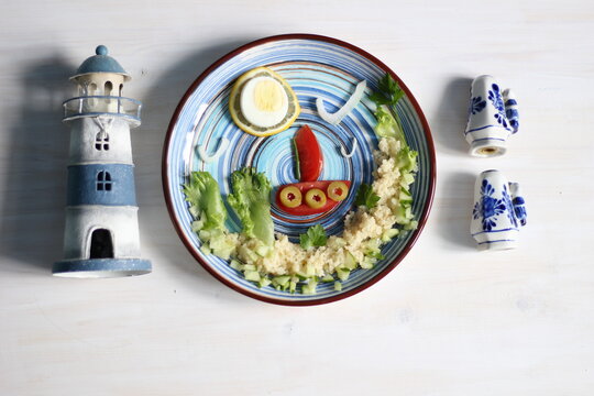 Salad with cuscus, vegetables, egg, created as picture of sea, boat, sun, palm trees in blue blate on white background with lighthouse