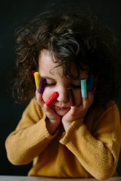 Portrait of sweet kid resting her head on her hands over dark background with marker caps on her fingers