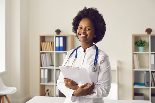 Portrait of happy professional doctor at work. Young black woman in white lab coat with stethoscope standing in medical office or exam room at clinic, holding clipboard, smiling and looking at camera