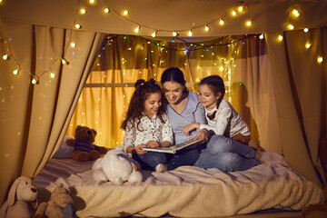 Obraz na płótnie Canvas Happy family reading good book in the evening. Young mother telling bedtime stories to little children. Mommy and daughters enjoying fairy tales sitting in cozy playroom tent decorated with LED lights