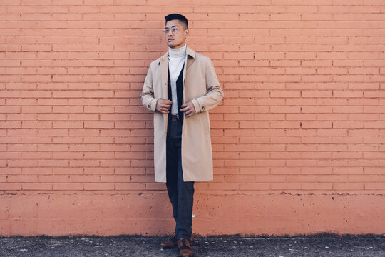 Middle-aged Latino man with glasses, beige trench coat, and brick background poses