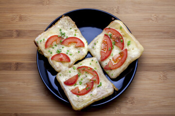 Egg-dipped toast bread and fried with melted cheese, tomato, and chives. Served on a shiny black plate lying on a wooden table.