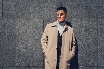 Middle-aged Latin man with glasses, beige trench coat, and gray background poses
