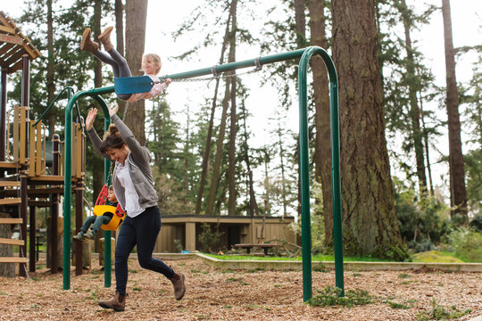 Playful Mom Pushing Her Kids On The Swings At The Park.