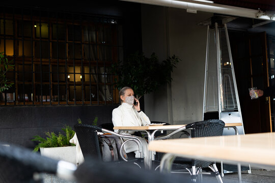 senior business woman talking by phone wearing mask while the waiter serves her a drink at restaurant