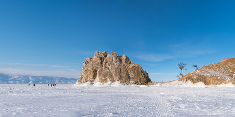Sacred Shamanka Mountain on Olkhon Island in winter. View from the frozen Lake Baikal. Tourists walk on the ice, admire the beauty of the ice. Panorama