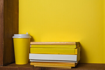On an old wooden shelf is a stack of old books with yellow covers. Nearby is a yellow paper cup for coffee