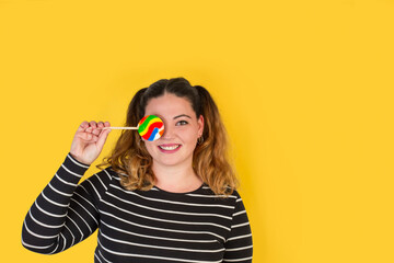 Mujer joven y sonriente sosteniendo un lollipop en la mano delante de un ojo sobre un fondo amarillo. Vista de frente y de cerca. Copy space