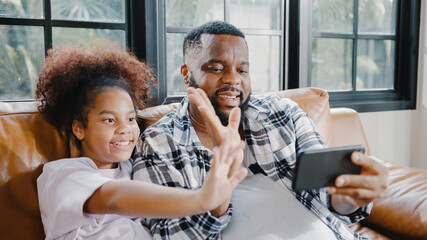 Happy African American family dad and daughter having fun and using mobile phone video call on sofa at house. Self-isolation, stay at home, social distancing, quarantine for coronavirus prevention.