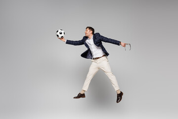 Fototapeta na wymiar full length of smiling businessman holding glasses and playing football while levitating on grey