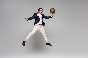 Fototapeta na wymiar full length of cheerful man in glasses playing basketball and levitating on grey