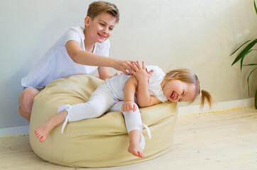Big brother and his cute little sister laying togetherness fun at home. Two Caucasian siblings brother and sister. Restless cheerful kids in casual clothes playing, having fun, hugging, smiling