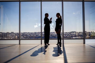 Two business women enjoying the city view and talking while standing by the large window in the office