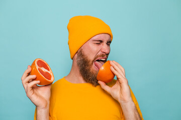 Bearded european man in yellow shirt isolated on turquoise background holding grapefruit tastes,...