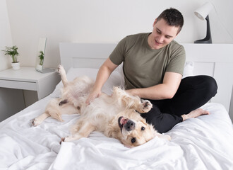 A young man is playing on the bed with a dog. Guy with companion golden retriever sitting at home