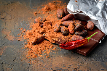 Dark chocolate, red chilli peppers, cocoa beans and powder on a dark stone background.