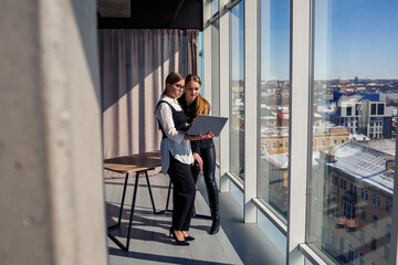 Two successful female architects are talking about a joint project while standing at the window with a laptop. Young women economists dressed in formal attire talking during a break from work