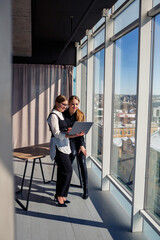 Two successful female architects are talking about a joint project while standing at the window with a laptop. Young women economists dressed in formal attire talking during a break from work