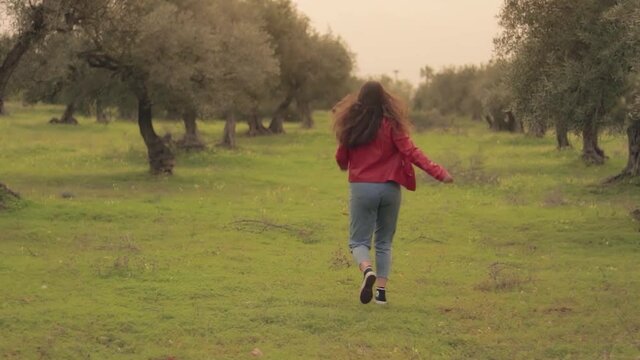 Mujer joven con chaqueta roja corriendo feliz en un prado verde con olivos y flores amarillas con el atardecer de fondo