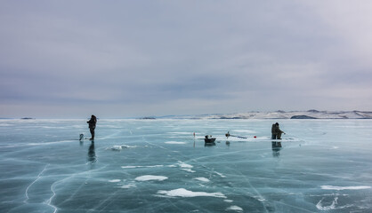Thick transparent ice with islands of snow is streaked with cracks. Two people are ice fishing. Next to them are fishing tackle, drill. In the distance, snow-covered hills. Cloudy. Russia. Lake Baikal