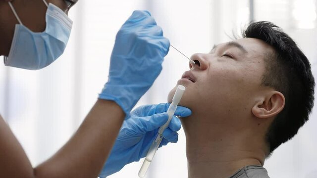 Side view of medical female worker in protective mask and gloves taking PCR test sample from asian man in hospital room. Concept of Covid reaesearch and diagnostic.