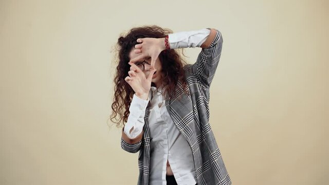 The curious young woman looks through her fingers to find out new things. Cretaceous in gray jacket and white shirt, with glasses posing isolated on a beige background in the studio. The concept of