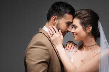 smiling bride touching face of muslim fiance while standing face to face isolated on grey