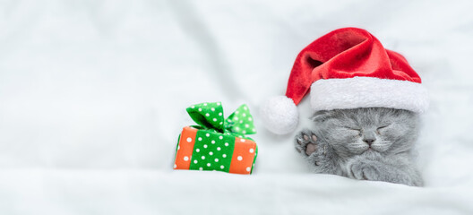 Cute kitten wearing red santa's hat sleeps with gift box under a white blanket on a bed. Top down...