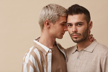 Minimal portrait of young gay couple embracing while posing against neutral beige background in studio, copy space