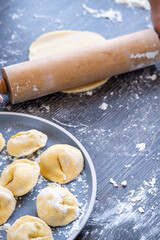 Pelmeni dumplings making process: Woman's hand flattening pelmeni dough using a rolling pin. Gluten free recipe for traditional Russian cuisine on a plate. Floured wooden surface, handmade dumplings.