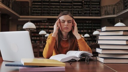 Portrait of tired female student in a yellow sweater and glasses sitting at a table in the university library read book feels a headache work on laptop using internet distance learning self isolation.