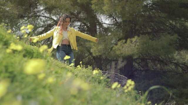 mujer joven con chaqueta amarilla bailando de felicidad en un bosque con un prado con flores amarillas en Espa&ntilde;a