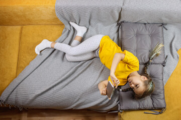 Digital native generation alpha, cute kid lying on yellow sofa with gray blanket and using smartphone for chatting and distant learning, online education at home, new normal lifestyle, trendy colors