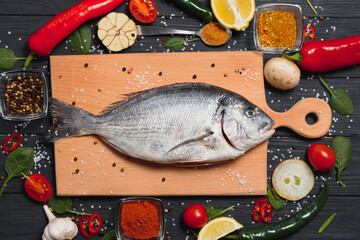 Fresh raw dorado fish on baking paper with lemon, pepper, tomatoes and various spices on wooden background with copy space.
