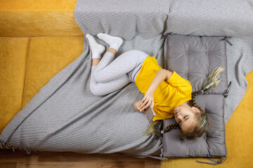 Digital native generation alpha, cute kid lying on yellow sofa with gray blanket and using smartphone for chatting and distant learning, online education at home, new normal lifestyle, trendy colors