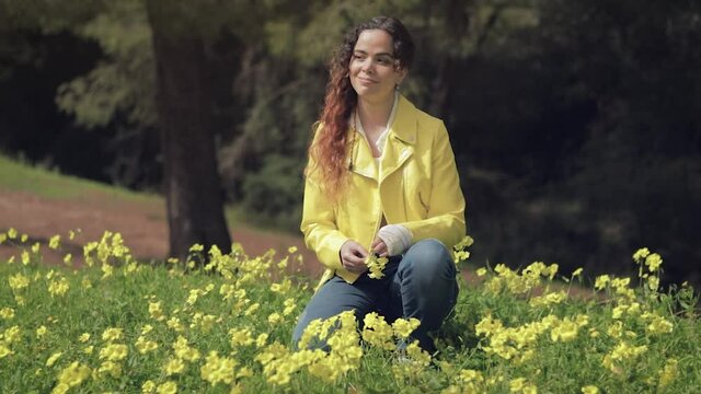 Mujer joven sonriendo y cubri&eacute;ndose los ojos del sol en un prado con flores amarillas en un bosque con &aacute;rboles