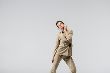 brunette woman in stylish pantsuit touching face while posing isolated on grey