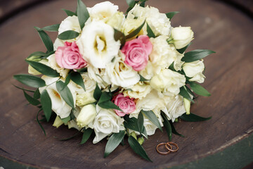 A wedding bouquet with lively white and pink roses lies on a wooden barrel near the gold wedding rings. Symbols of eternal love on the table. Natural bouquet of beautiful flowers. 