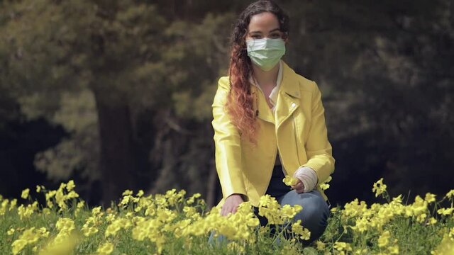 Mujer joven con mascarilla quir&uacute;rgica arrodillada en un prado con flores amarillas en un bosque con &aacute;rboles