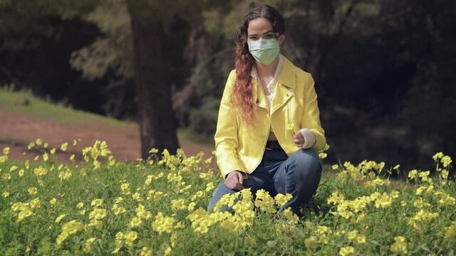 Mujer joven con mascarilla quir&uacute;rgica arrodillada en un prado con flores amarillas en un bosque con &aacute;rboles