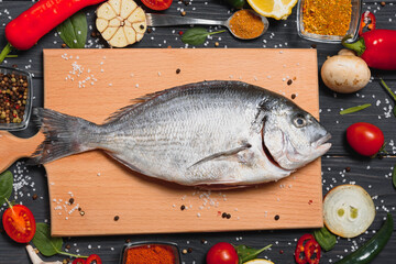 Fresh raw dorado fish on baking paper with lemon, pepper, tomatoes and various spices on wooden background with copy space.