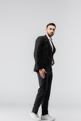 Obraz na płótnie Canvas young arabian man in elegant suit and sneakers looking at camera while posing on grey