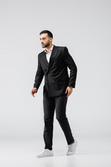 Obraz na płótnie Canvas trendy muslim man in black suit and sneakers looking away while walking on grey