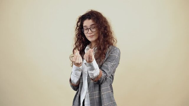 The beautiful employee shows with her hand the gesture of money, in order to receive a payment bonus. Cretaceous in gray jacket and white shirt, with glasses posing isolated on a beige background in