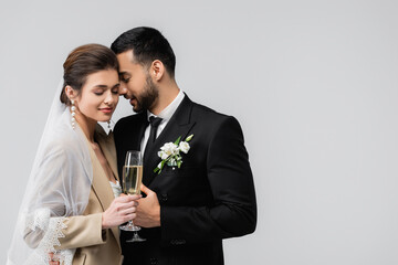 joyful, interracial newlywed couple standing with closed eyes and champagne glasses isolated on grey