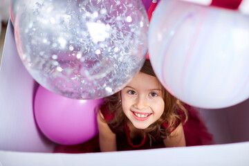 Cute, joyful little girl in pink dress looks out of the big present gift box with balloons